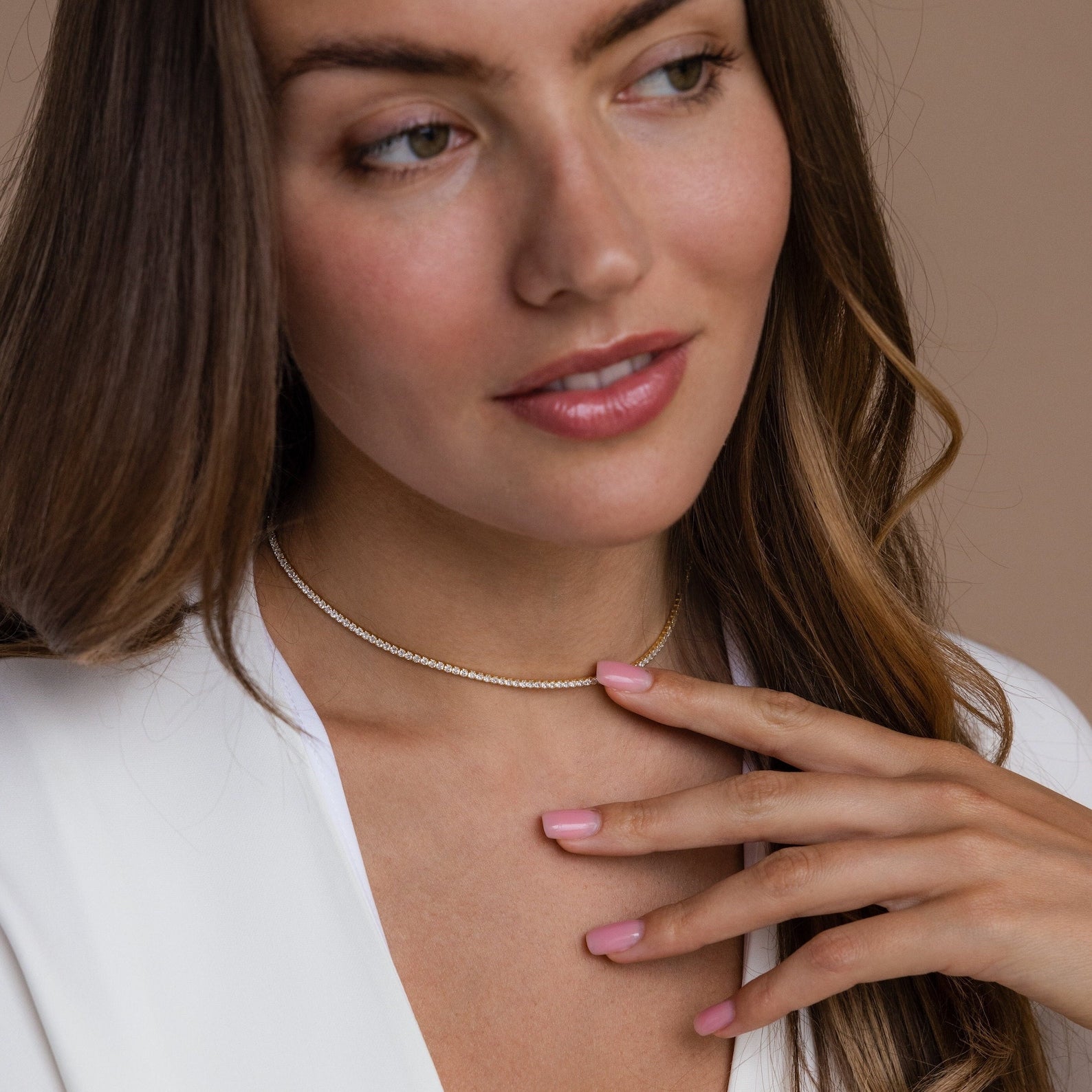 A woman in a white top softly touching her gold diamond tennis choker necklace, with a serene expression and pink manicured nails.