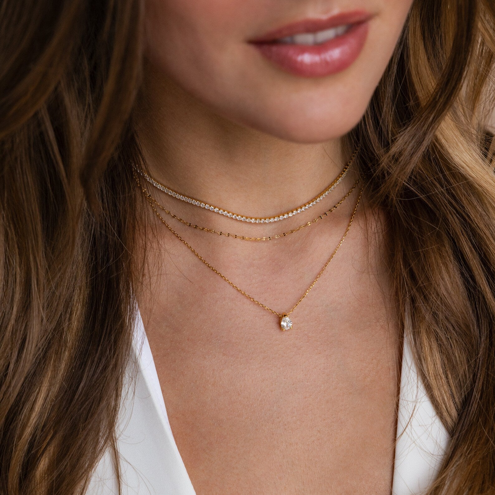A layered necklace look featuring a gold diamond tennis choker paired with two gold chain necklaces and a teardrop pendant, worn over a white blouse.
