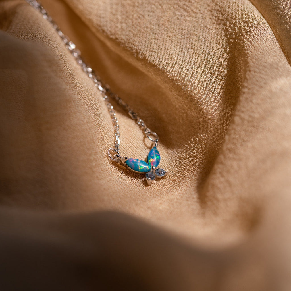 The Blue Opal Butterfly Necklace features a delicate butterfly pendant, shown on soft beige fabric in gentle light—a dainty accessory perfect for any occasion.