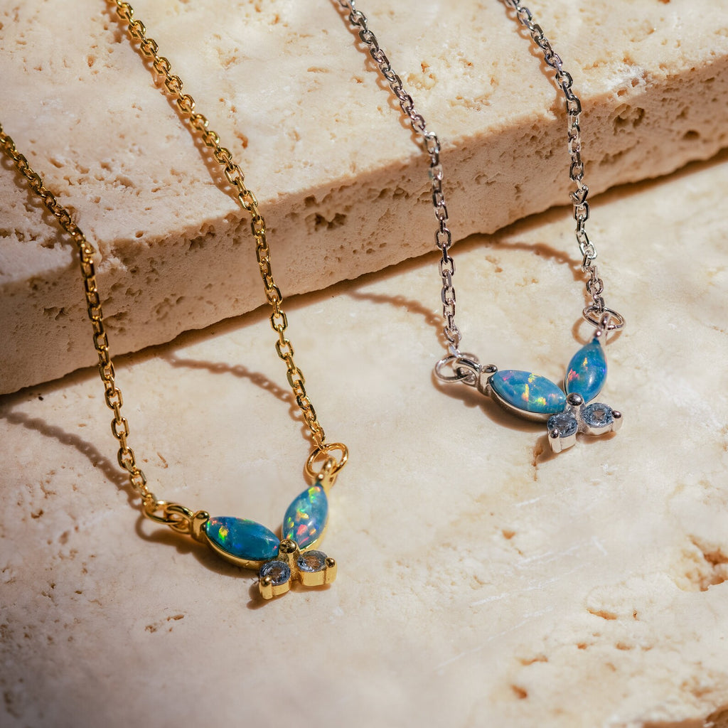 Gold and silver butterfly necklaces with blue opal wings and clear stone accents, displayed on a textured stone surface.
