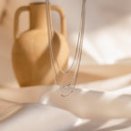 The Triple Box Chain Necklace, a delicate silver layered piece, is displayed hanging in front of a ceramic vase on silky cream-colored fabric.