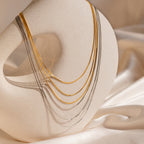 The Triple Box Chain Necklace in gold and silver drapes elegantly over a sculptural beige stand on silky fabric.