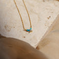 The Blue Opal Butterfly Necklace features a gold chain and a blue opal butterfly pendant, displayed on a textured beige stone surface. This dainty necklace adds elegance to any outfit.