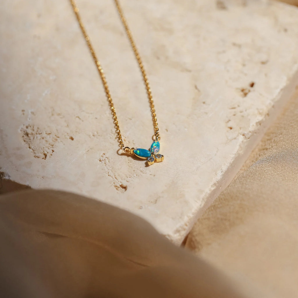Opal Butterfly Necklace Set with a gold chain and delicate blue opal butterfly pendant, displayed on a light textured stone surface.