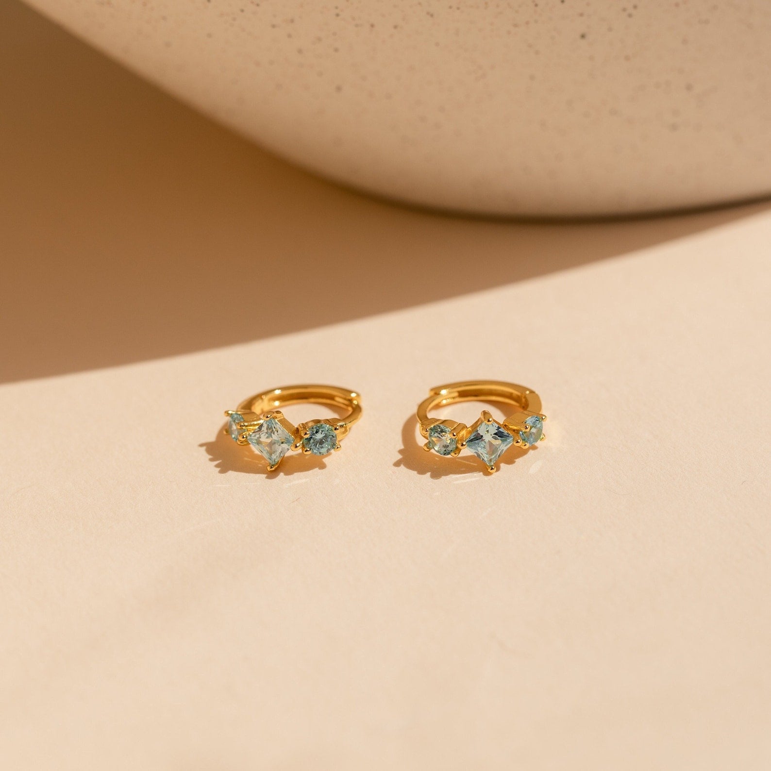 The gold Aquamarine Diamond Huggies displayed flat with their aquamarine stones facing forward on a warm beige background.