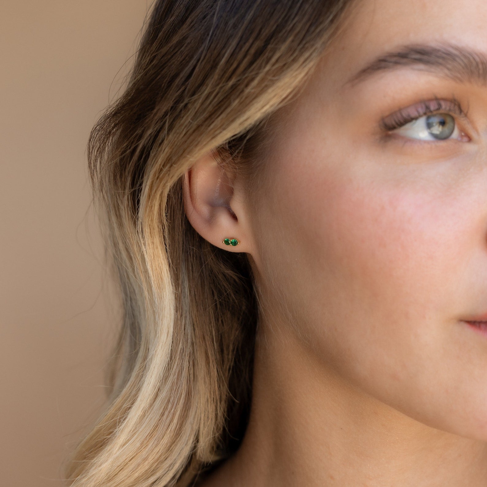 Close-up of a woman's ear wearing Duo Birthstone Studs with green gemstones; part of her face visible—an ideal, thoughtful bridesmaids gift.