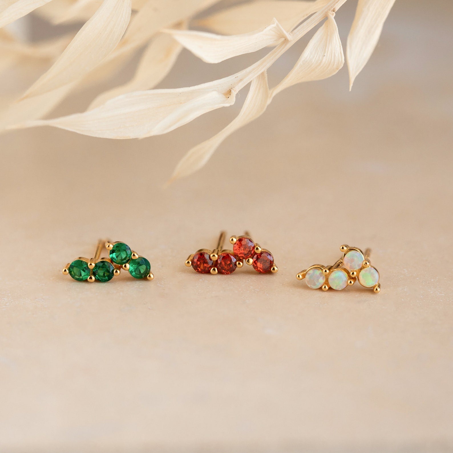 Three pairs of Duo Birthstone Studs in gold, featuring green, red, and opal gemstones, displayed on a neutral background—an ideal bridesmaids gift.