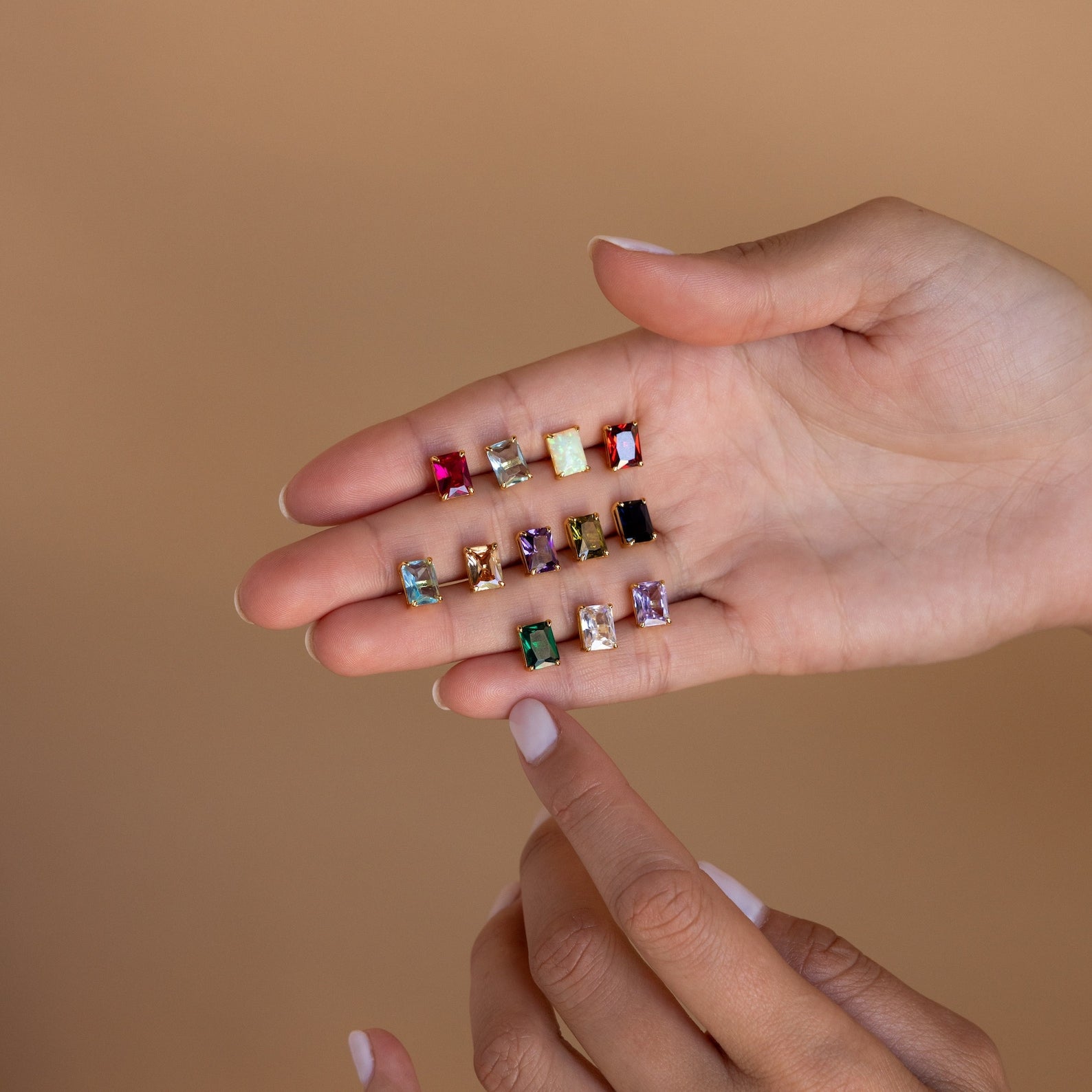 A hand showcases Emerald Birthstone Studs, perfect as personalized gifts, while another hand points to one of the sparkling earrings.