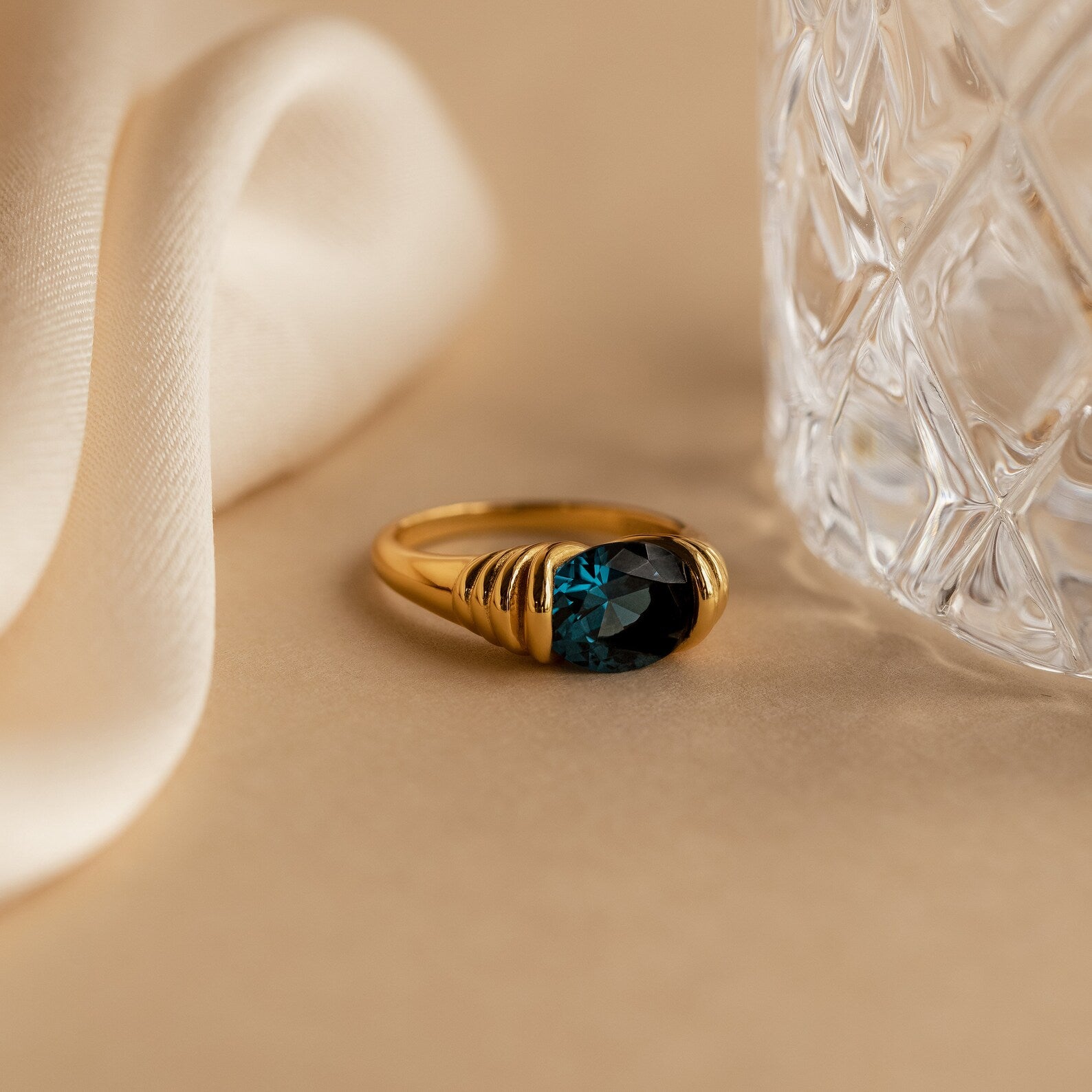 The London Blue Signet Ring features a striking London Blue Topaz gemstone, showcased beside a crystal glass and set against a beige fabric backdrop.