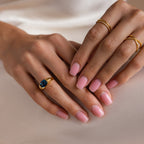 Hands with light pink manicured nails wear gold rings, including the London Blue Signet Ring, resting on a white surface.