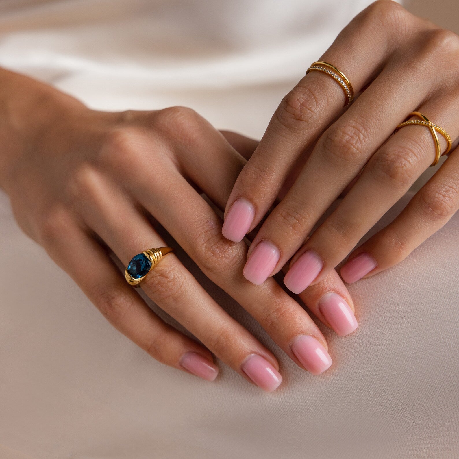 Hands with light pink manicured nails wear gold rings, including the London Blue Signet Ring, resting on a white surface.