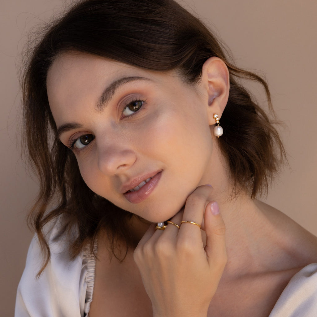 Front-facing image of a model softly smiling while wearing gold Millie Drop Earrings, styled with minimal makeup and gold rings.