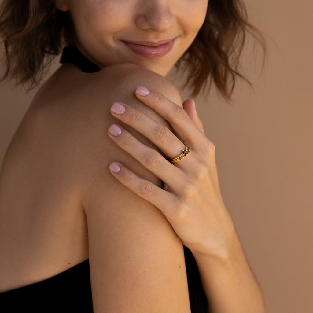A woman with light pink nails smiles, showing off the Black Abyss Ring Set—dainty bands with black gemstones—on her finger, her bare shoulder against a beige background.
