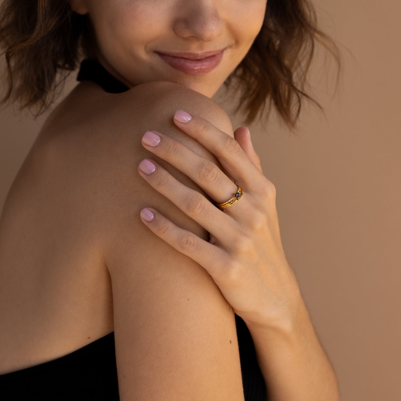 A woman with light pink nails smiles, showing off the Black Abyss Ring Set—dainty bands with black gemstones—on her finger, her bare shoulder against a beige background.