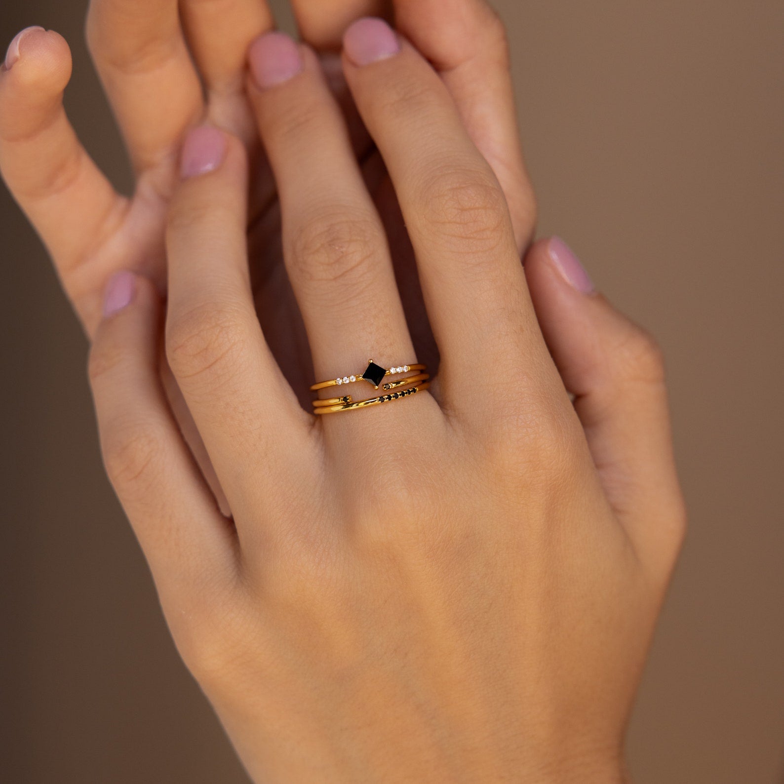 A hand with pink nails wears three stacked gold rings, including the Black Abyss Ring Set, which features delicate bands and a small black geometric stone.