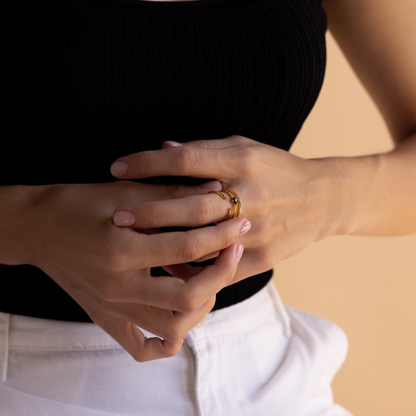 A woman in a black top and white pants wears the Black Abyss Ring Set—delicate bands featuring black gemstones—with her hands clasped at her waist.
