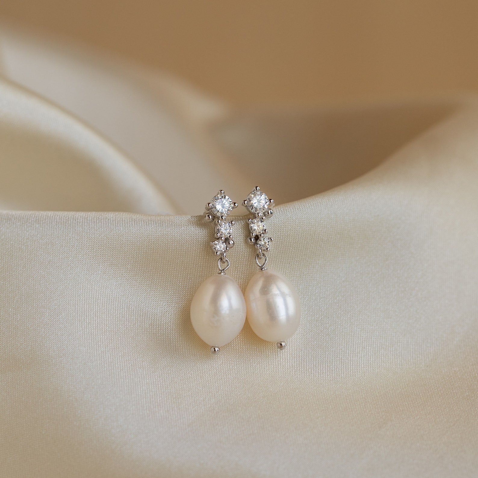 Silver Millie Drop Earrings displayed on satin fabric, with round-cut stones and freshwater pearls in soft lighting.