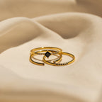Three gold rings with small black gemstones from the Black Abyss Ring Set displayed on soft beige fabric.