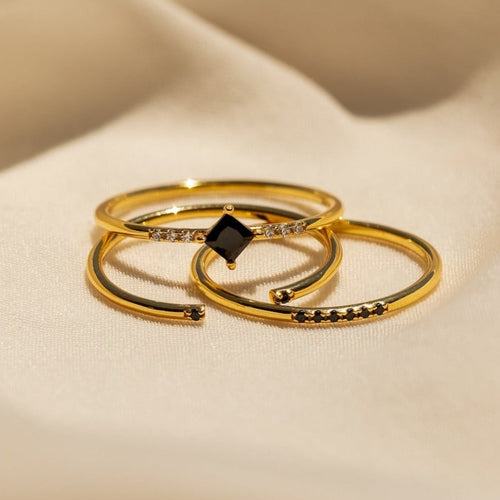 Three gold rings with small black gemstones from the Black Abyss Ring Set displayed on soft beige fabric.