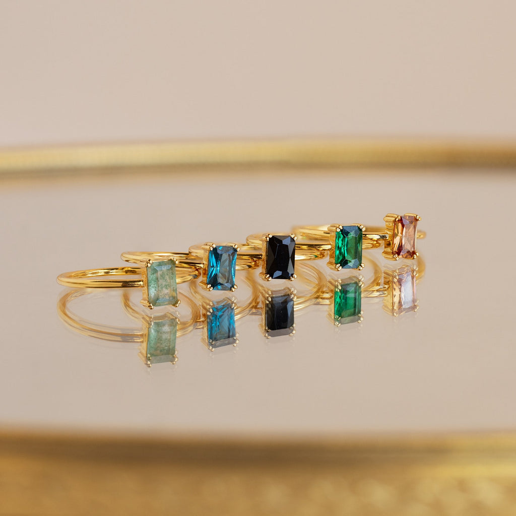 A Blue Topaz Emerald Ring shines among six gold rings with colorful rectangular gemstones, all displayed on a reflective surface that beautifully enhances their vibrant reflections.