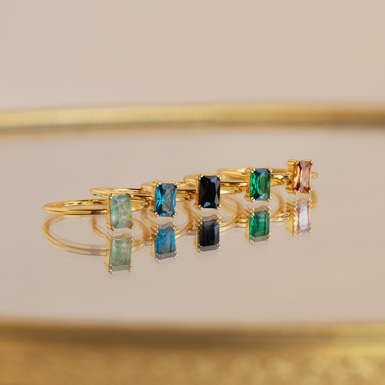 A Blue Topaz Emerald Ring shines among six gold rings with colorful rectangular gemstones, all displayed on a reflective surface that beautifully enhances their vibrant reflections.