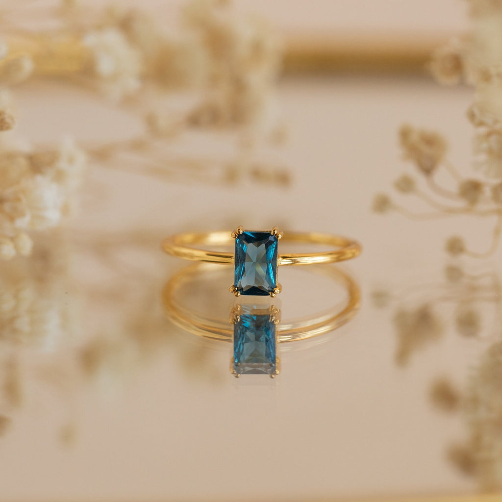 The Blue Topaz Emerald Ring is a vintage gold accessory featuring a rectangular blue topaz, beautifully reflected on a surface and framed by softly blurred flowers.