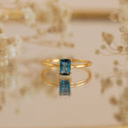 The Blue Topaz Emerald Ring is a vintage gold accessory featuring a rectangular blue topaz, beautifully reflected on a surface and framed by softly blurred flowers.