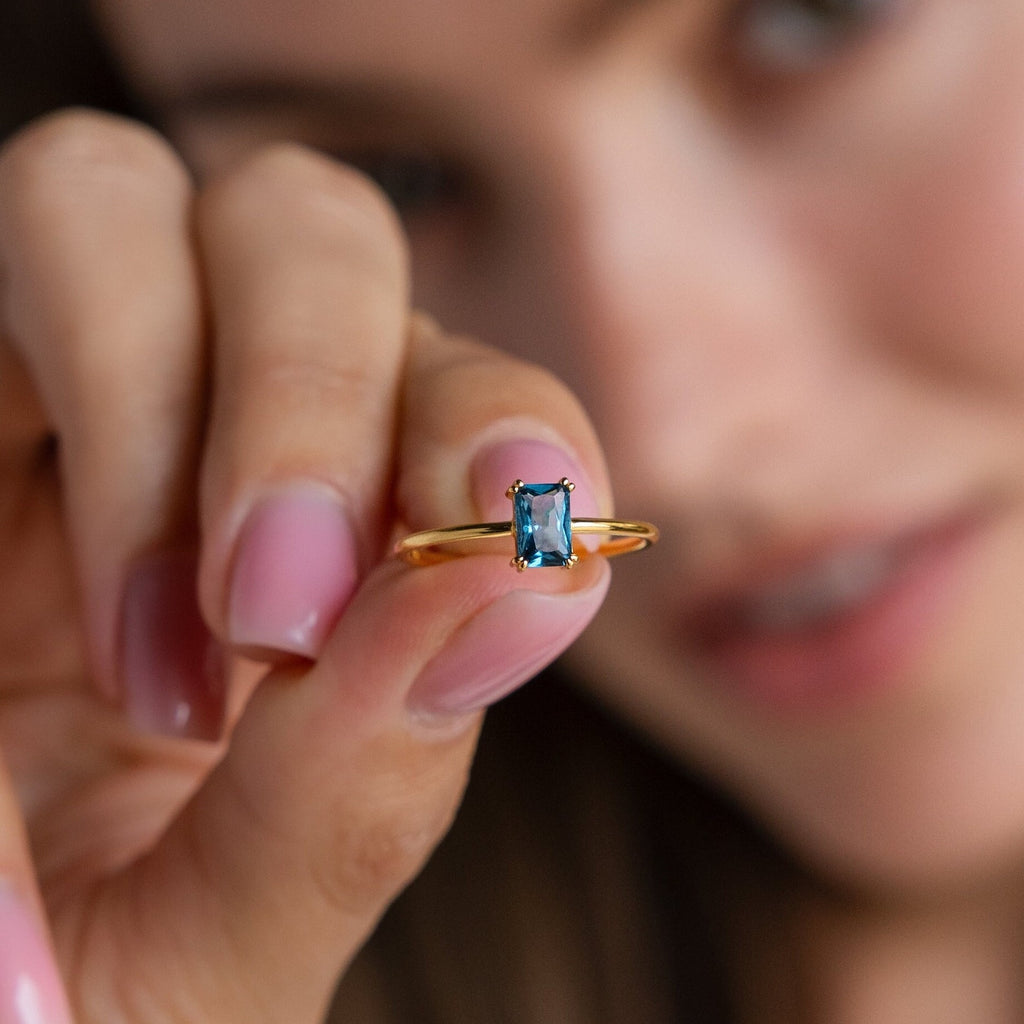 A woman with pink nails holds a gold Blue Topaz Emerald Ring featuring a striking rectangular gemstone close to the camera, beautifully showcasing this vintage accessory.