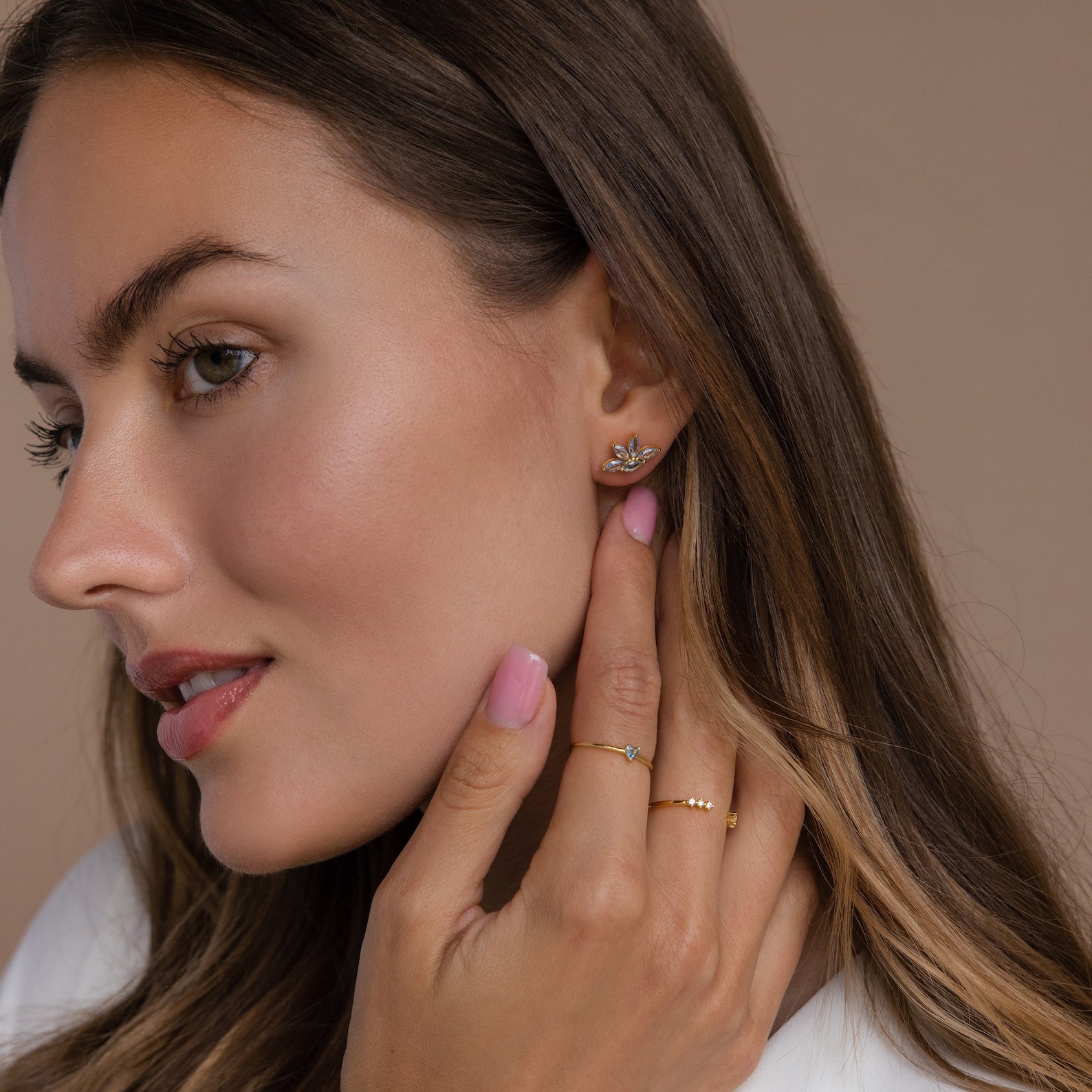 Woman with long brown hair wears Aquamarine Marquise Studs and gold rings, gently touching her face—these chic, floral-inspired earrings make a perfect accessory or thoughtful bridesmaid gift.