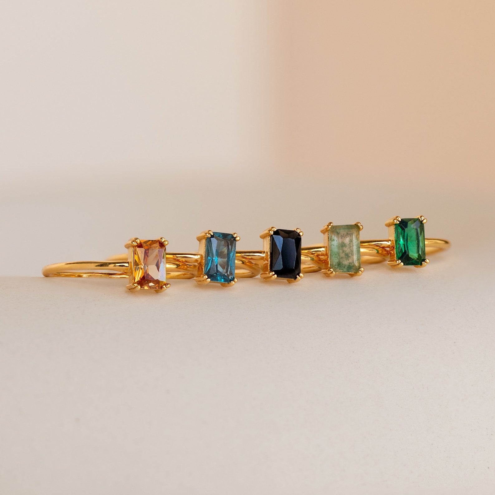 A row of five gold rings, including the Blue Topaz Emerald Ring with rectangular gemstones in vibrant hues, are arranged on a soft beige surface—an ideal vintage-inspired accessory collection.