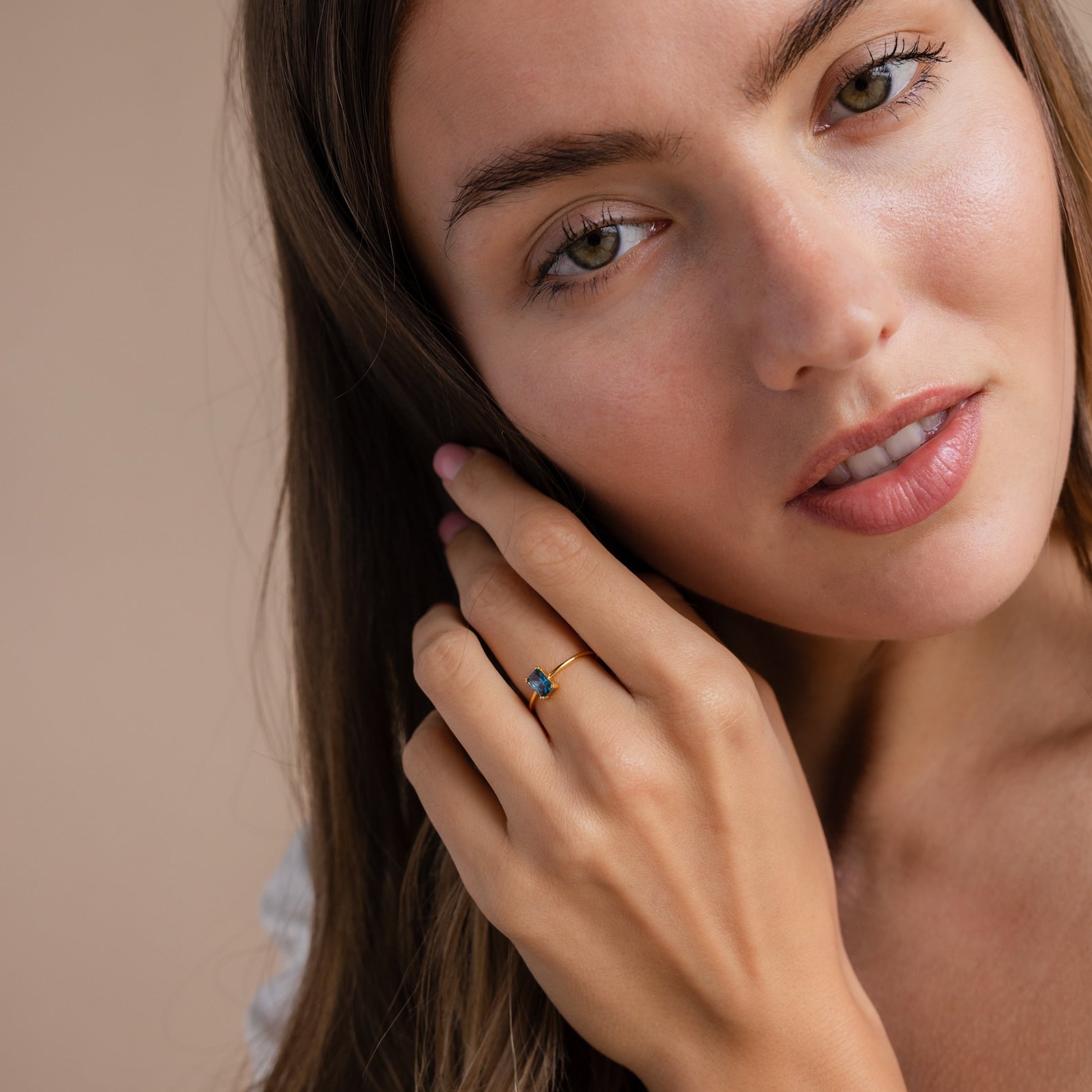 A woman with long brown hair showcases the Blue Topaz Emerald Ring, featuring a gold band, as she looks at the camera—a stunning vintage-inspired accessory.