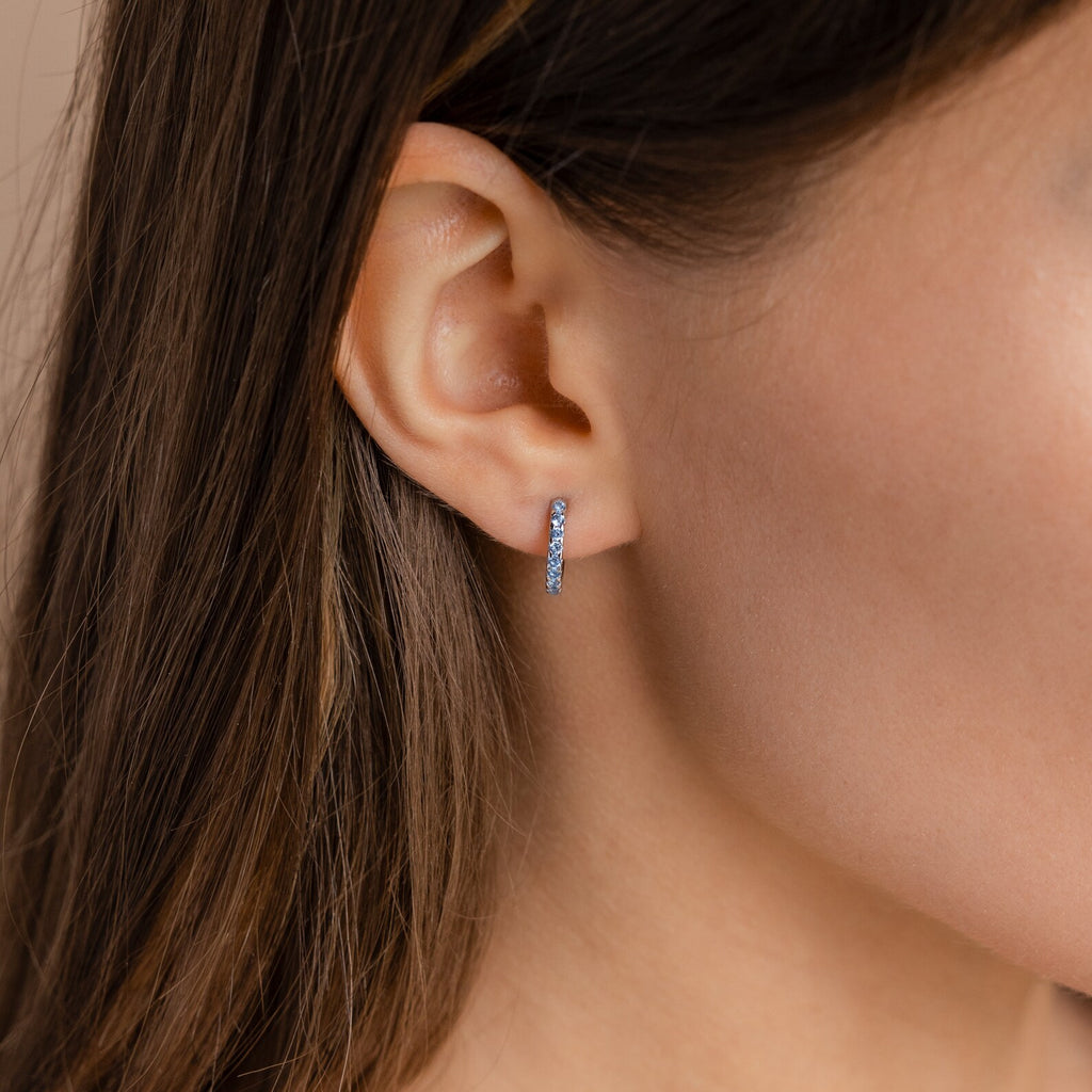 Close-up of a woman’s ear wearing the Aquamarine Pave Huggies—small, silver hoop earrings with embedded stones—styled as part of an elegant earring stack.