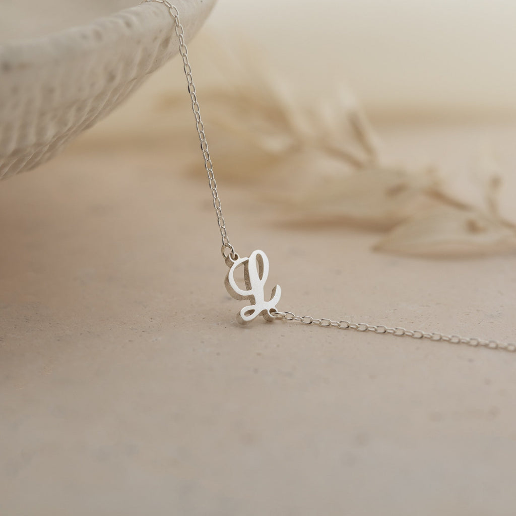 Delicate silver necklace with a cursive letter L pendant on a neutral surface—perfect for pairing with your favorite personalized jewelry or the Carrie Initial Bracelet.