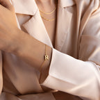 A woman wearing a gold Newsletter Initial Bracelet and layered gold necklaces, styled with a beige blazer.