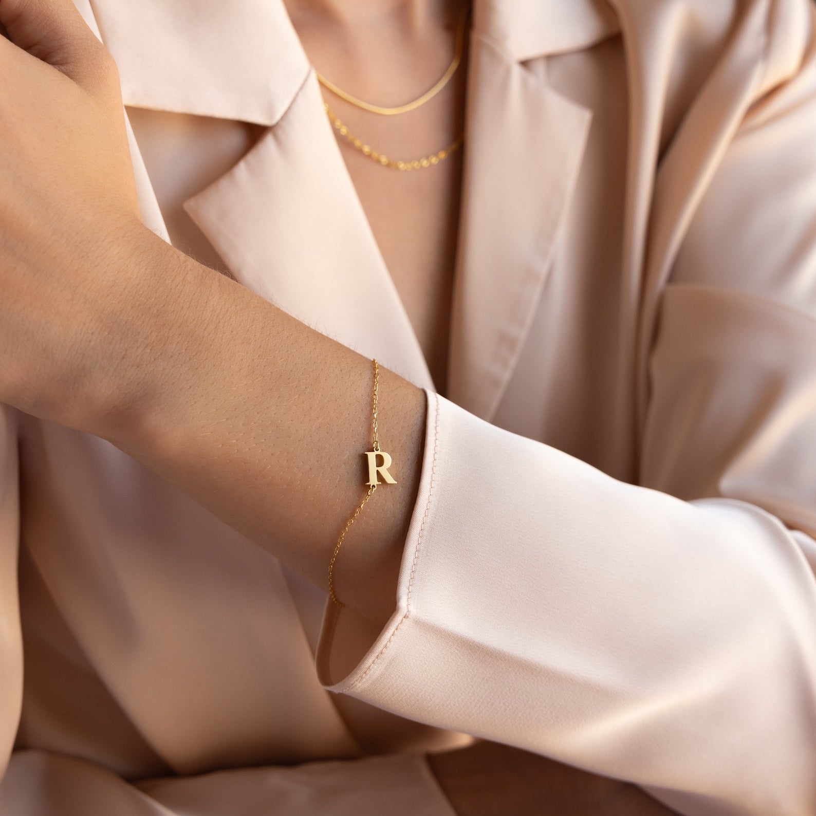 A woman wearing a gold Newsletter Initial Bracelet and layered gold necklaces, styled with a beige blazer.