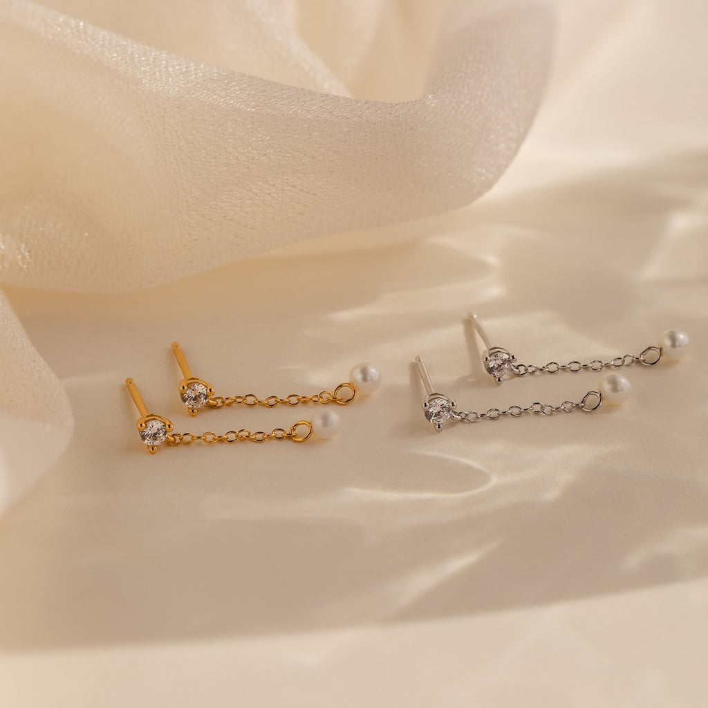 Diamond Pearl Drop Earrings featuring gold and silver accents with pearls and crystals are displayed on a soft, cream-colored fabric background—an elegant accessory to enhance any outfit.