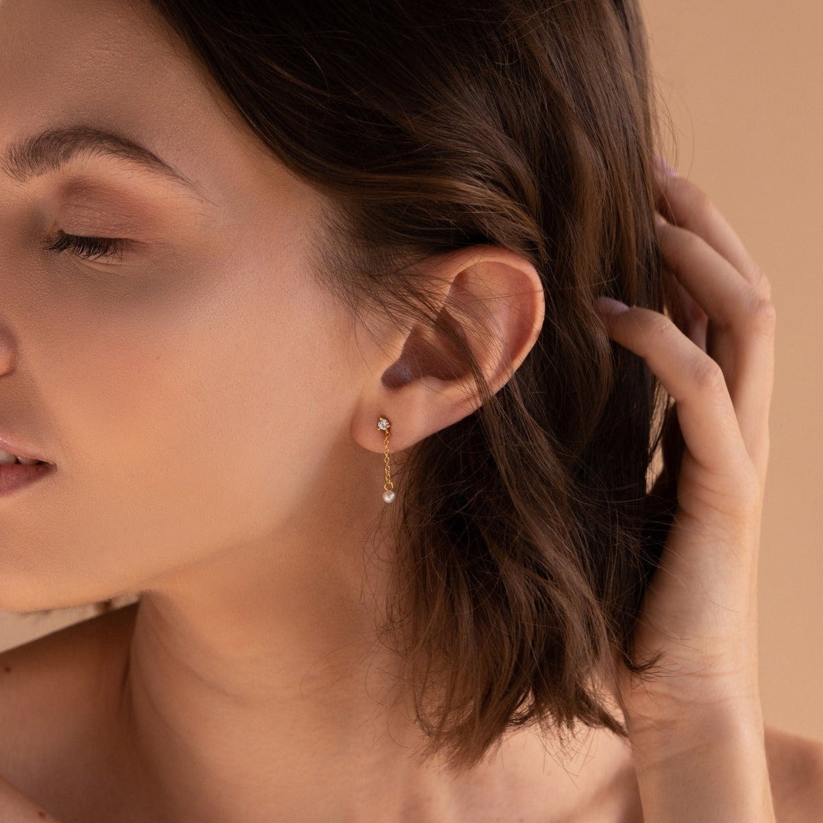 A woman with short brown hair touches her ear, displaying Diamond Pearl Drop Earrings—a delicate gold design featuring a small dangling gem.