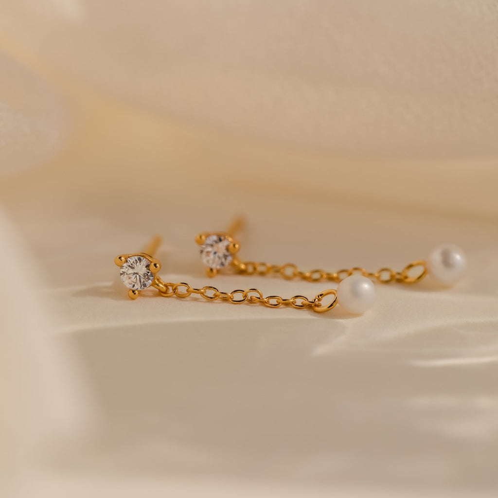 Diamond Pearl Drop Earrings feature sparkling diamonds and pearls on gold chains, beautifully displayed against a soft beige fabric background.