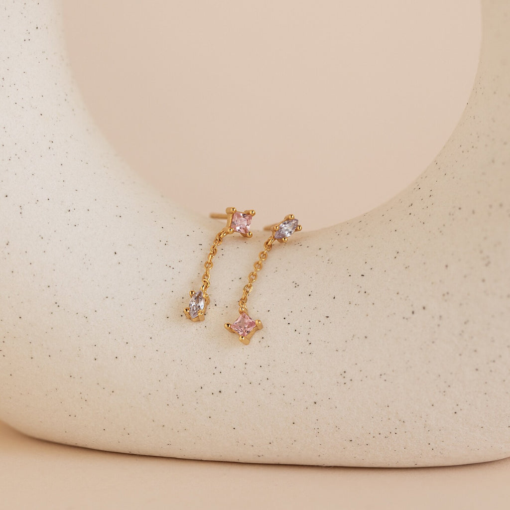 Mixed Crystal Drop Earrings featuring gold chains with pink and clear gemstones are displayed on a curved, speckled beige ceramic surface, adding subtle elegance to any look.