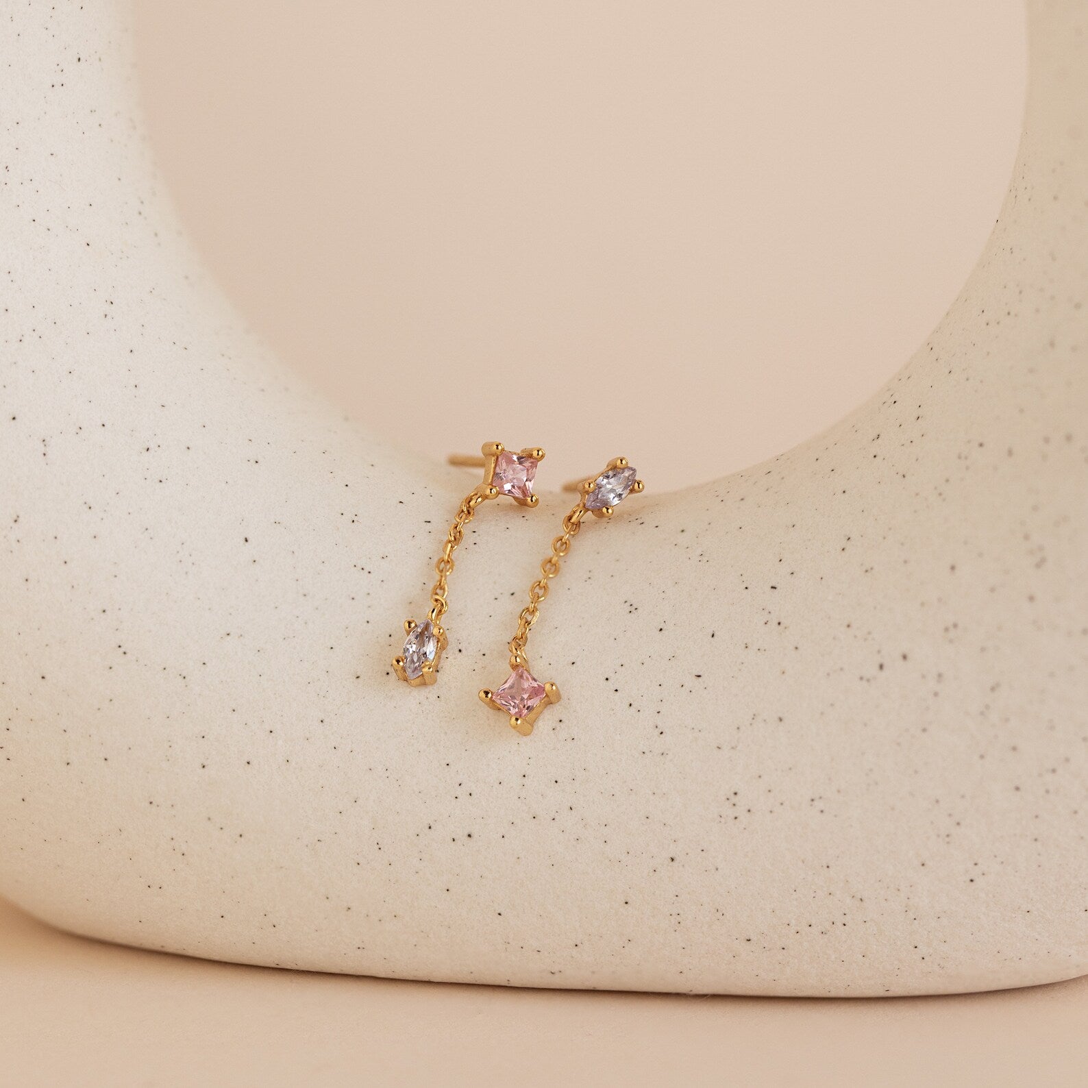Mixed Crystal Drop Earrings featuring gold chains with pink and clear gemstones are displayed on a curved, speckled beige ceramic surface, adding subtle elegance to any look.