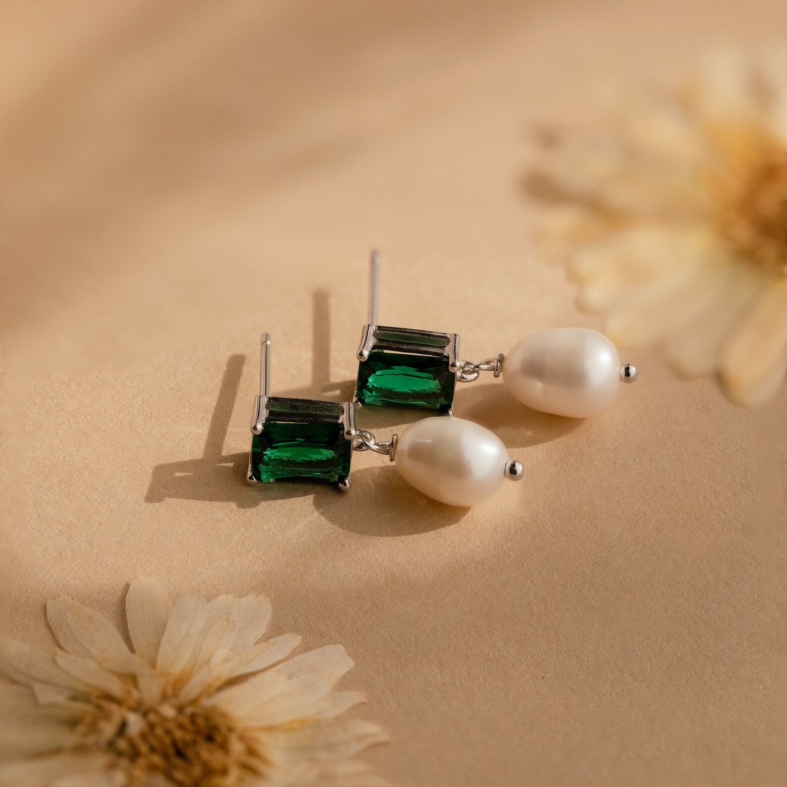 Emerald Pearl Drop Earrings featuring vintage emerald green gemstones, displayed on a beige surface with dried flowers.