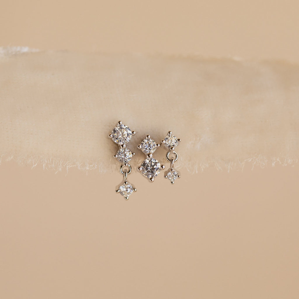 The Triple Diamond Studs Set features delicate triple diamond studs with a small dangling diamond, perfect for stacking earrings, displayed on a beige textured surface.