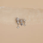 The Triple Diamond Studs Set features delicate triple diamond studs with a small dangling diamond, perfect for stacking earrings, displayed on a beige textured surface.