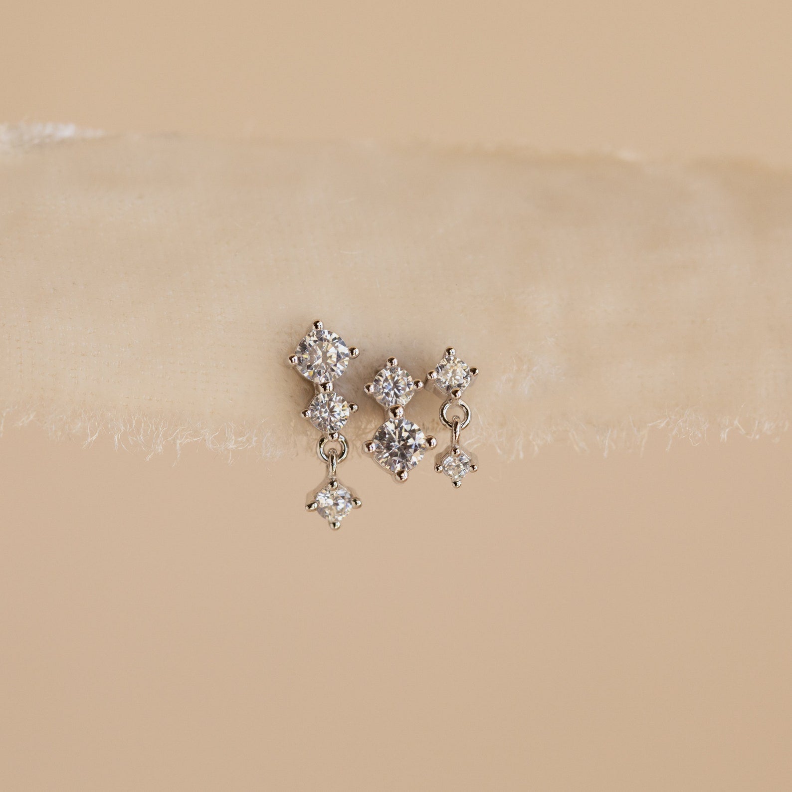 The Triple Diamond Studs Set features delicate triple diamond studs with a small dangling diamond, perfect for stacking earrings, displayed on a beige textured surface.
