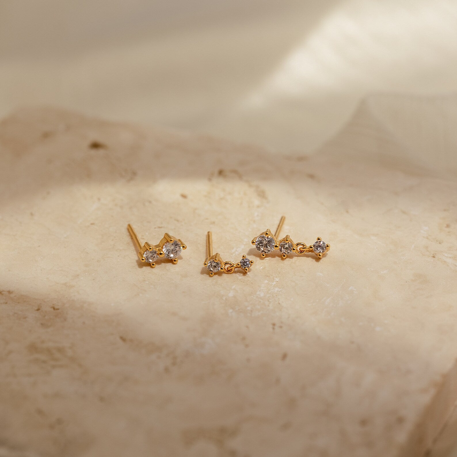 Triple Diamond Studs Set featuring small sparkling stones, presented on a textured beige stone surface in soft lighting.