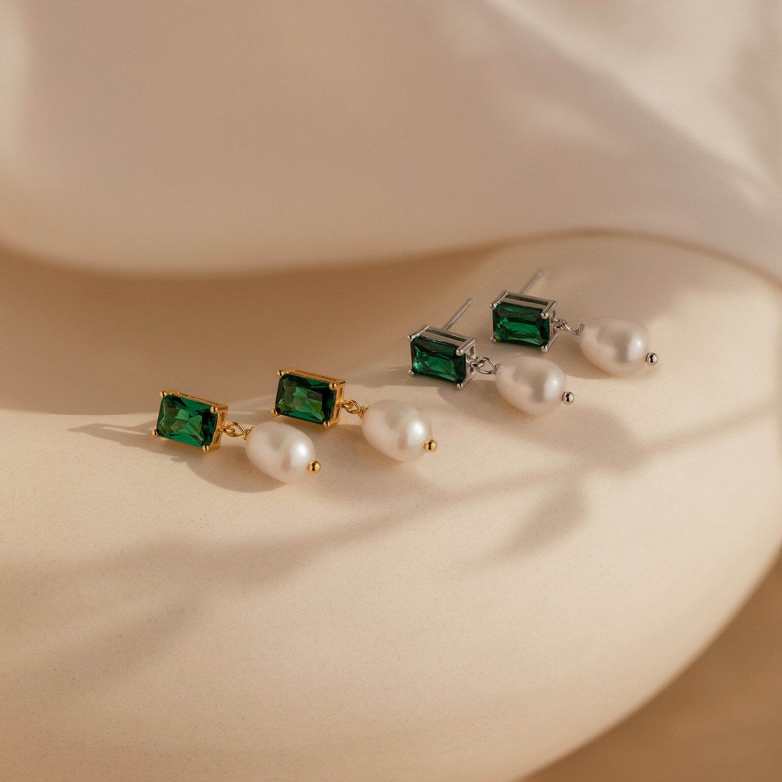 The Emerald Pearl Drop Earrings, featuring both gold and silver styles with green gemstones and pearls, are displayed on a neutral background.