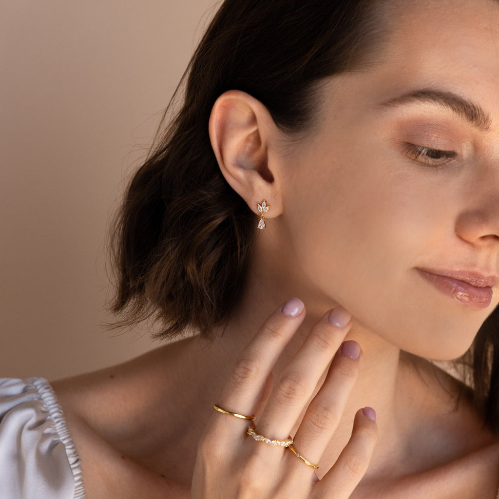A woman with short brown hair touches her face against a neutral background, wearing Marquise Diamond Drop Studs and gold rings.
