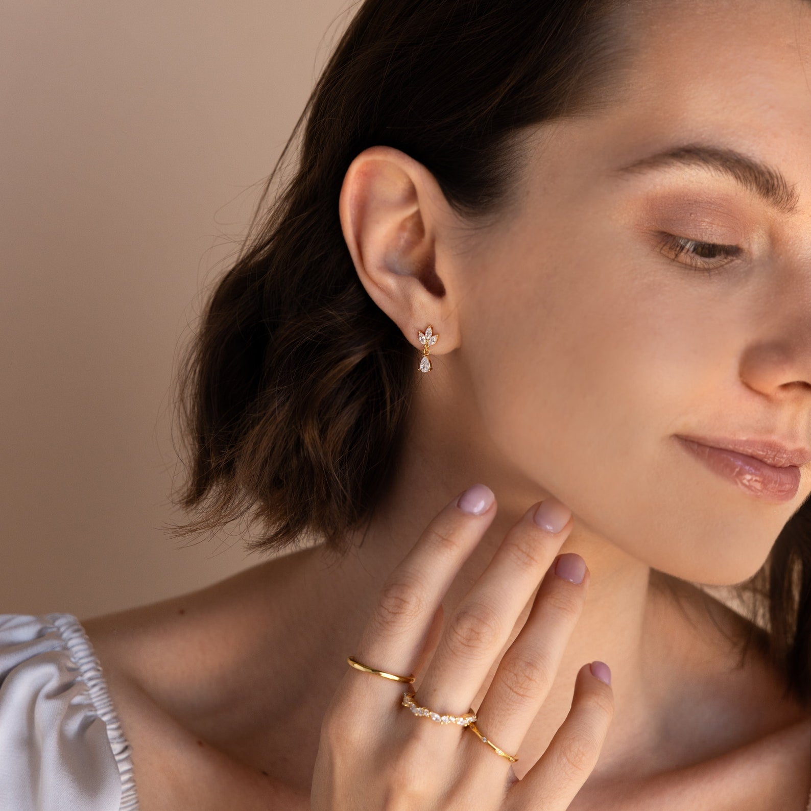 A woman with short brown hair touches her face against a neutral background, wearing Marquise Diamond Drop Studs and gold rings.