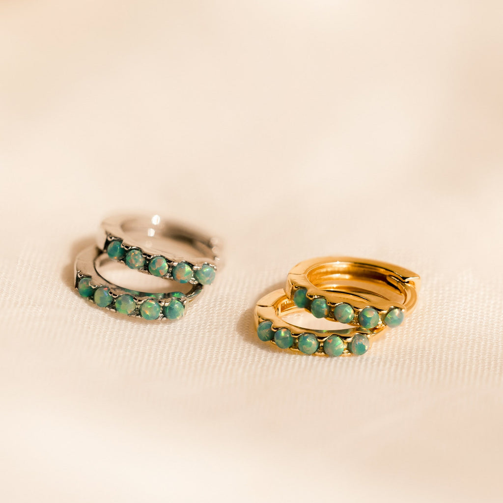 Two pairs of Garden Green Opal Huggies—one silver, one gold—featuring green gemstone accents for a chic earring stack, are displayed on a soft beige surface.