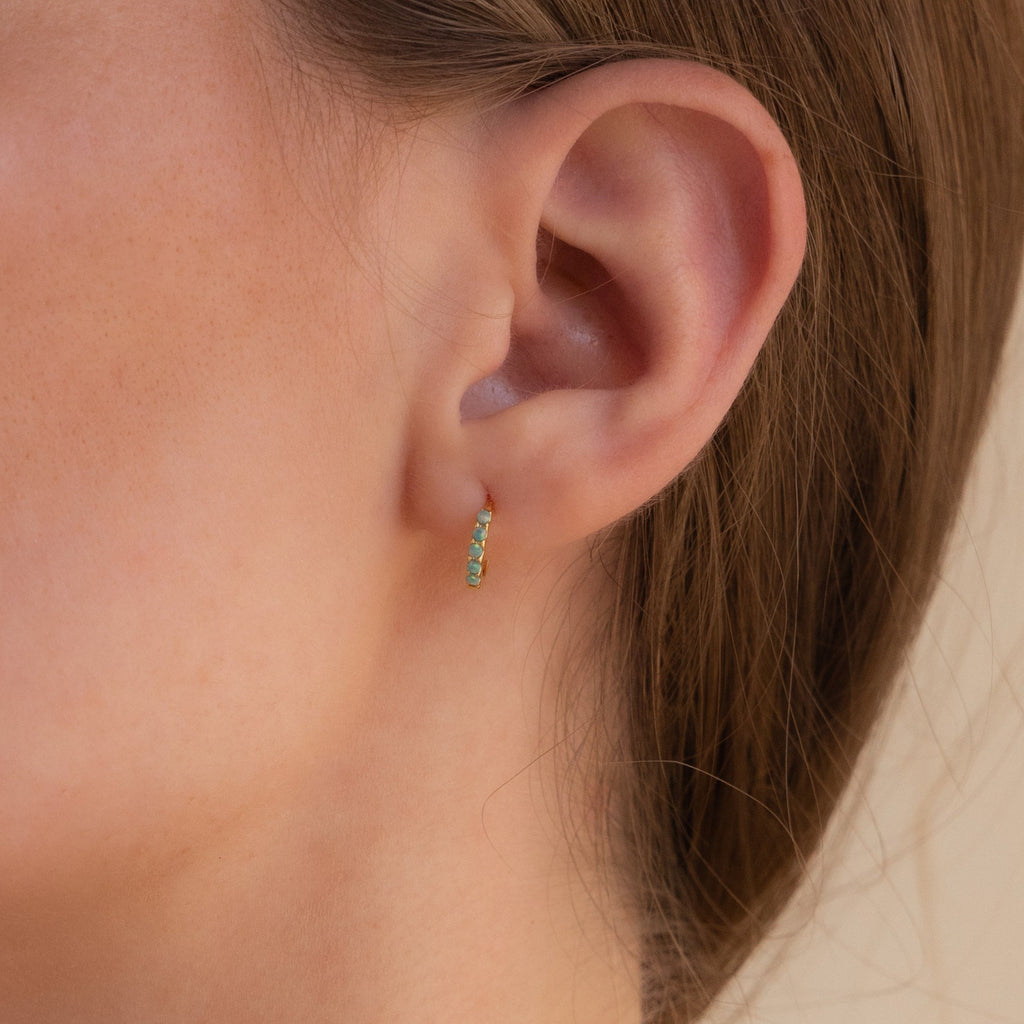 A close-up of a woman's ear adorned with an elegant earring stack, featuring the Garden Green Opal Huggies and green gemstone hoop earrings.